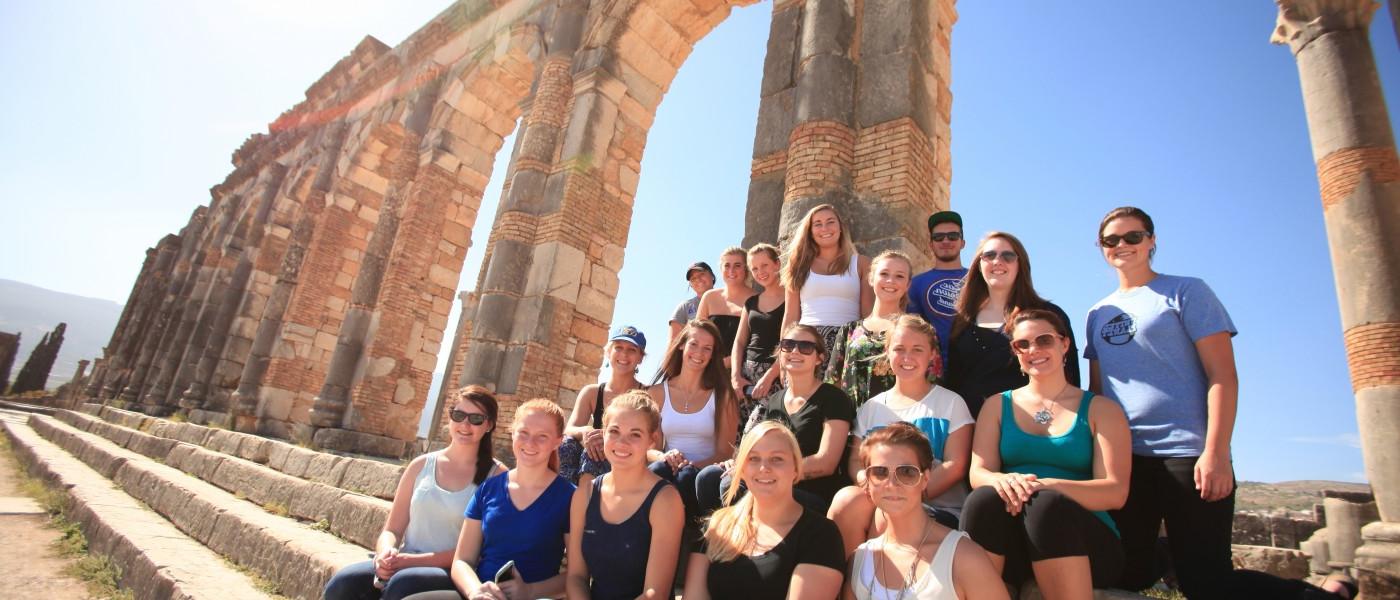 A group of U N E students sit on the steps by column ruins in Morocco